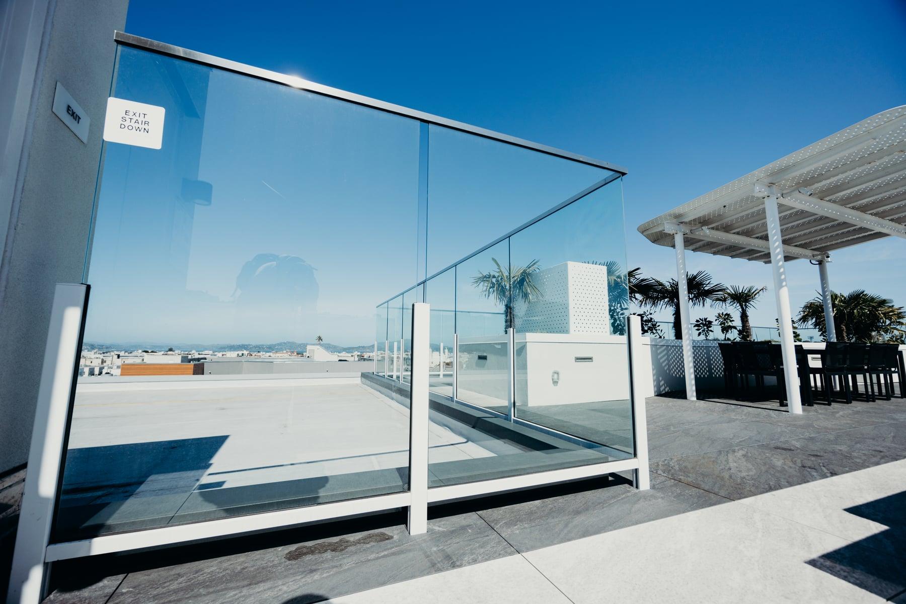 Glass guardrail around a rooftop terrace with a white pergola and palm trees under blue sky