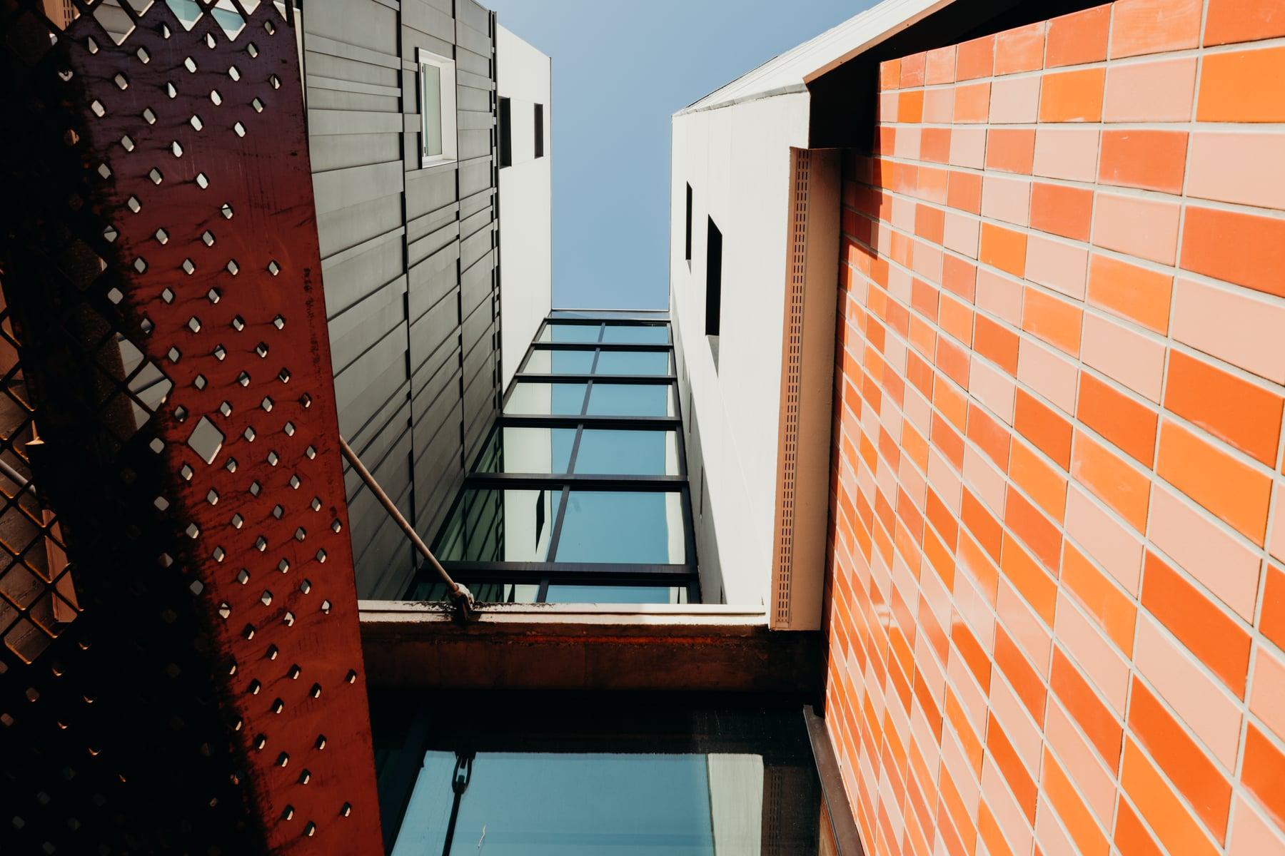 Looking up between two sections of the building: orange tile cladding, a dark perforated metal panel and a glazed passage between them