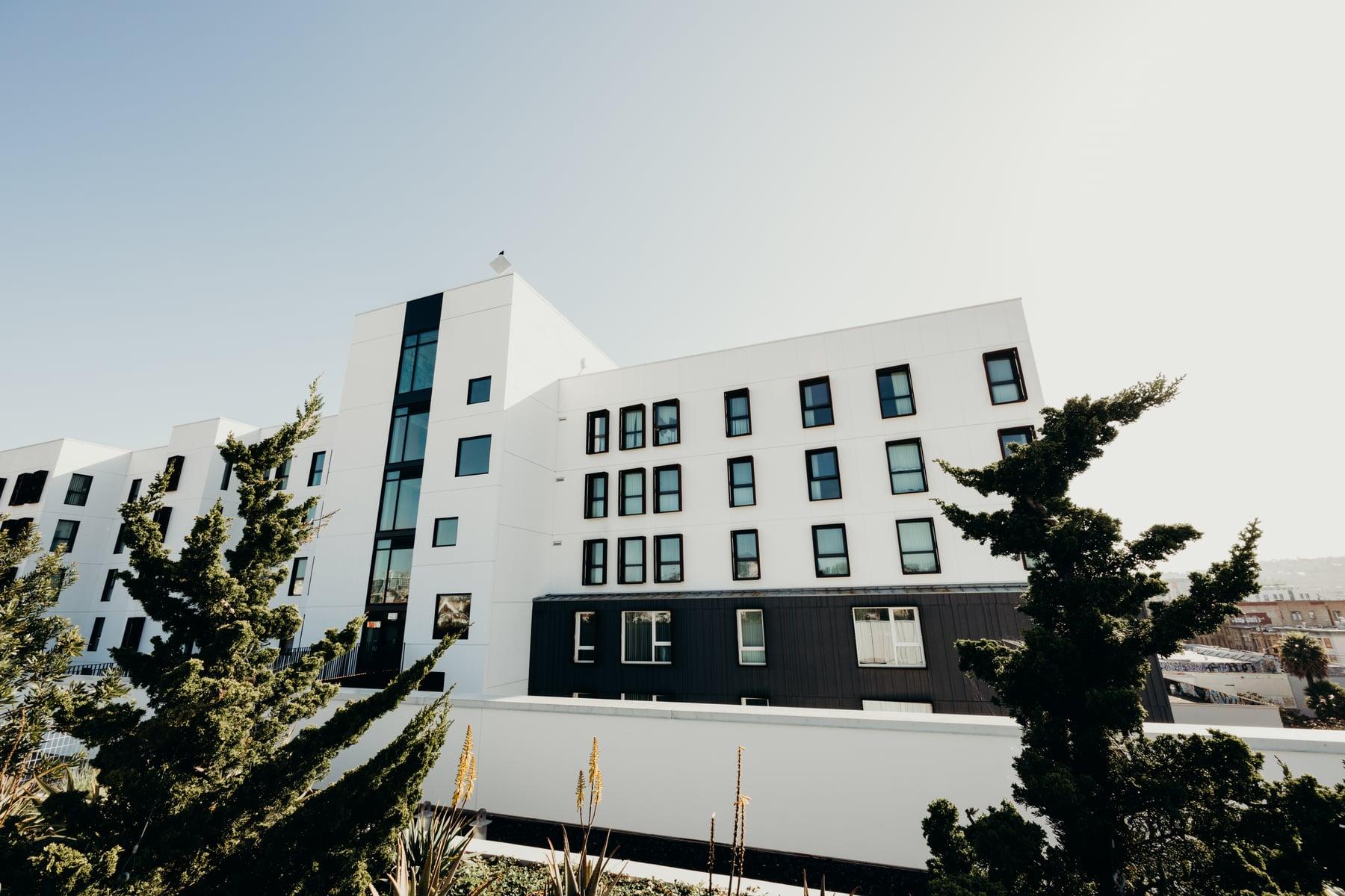 White and dark-paneled residential building with rows of windows behind trees