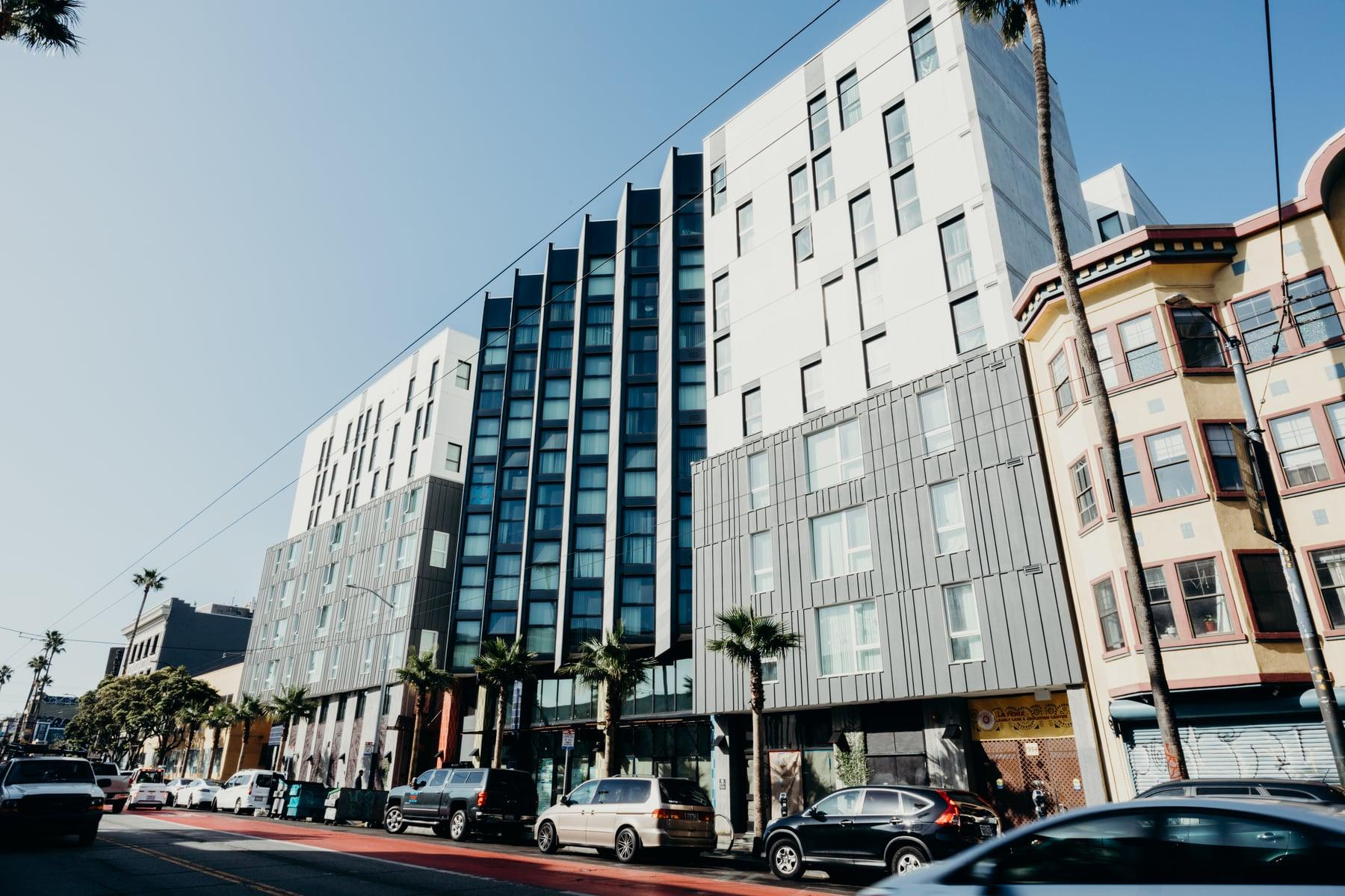 Street view of the building's white, gray metal-panel and dark blue glazed sections beside older buildings, with parked cars