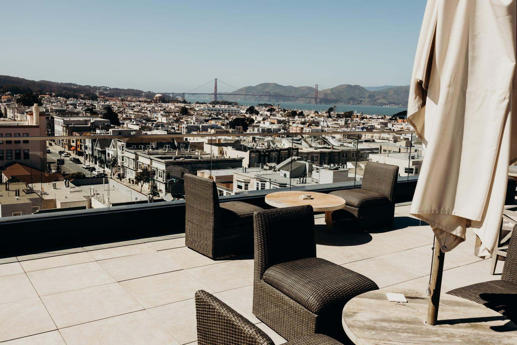 Rooftop terrace with wicker seating and an umbrella, overlooking San Francisco rooftops toward a red suspension bridge
