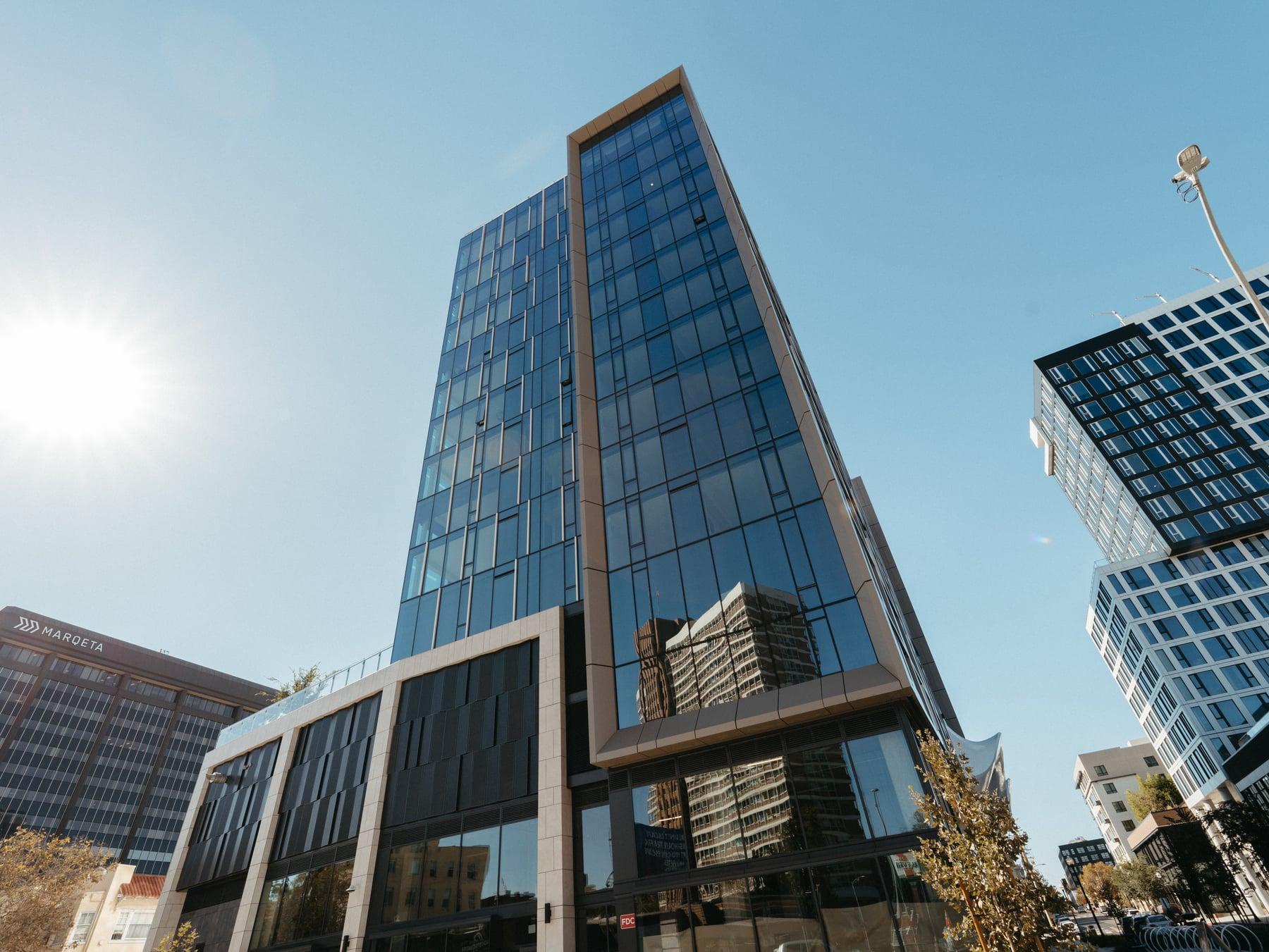 Looking up at a glass-clad tower with angled blue-tinted glazing against a sunny sky