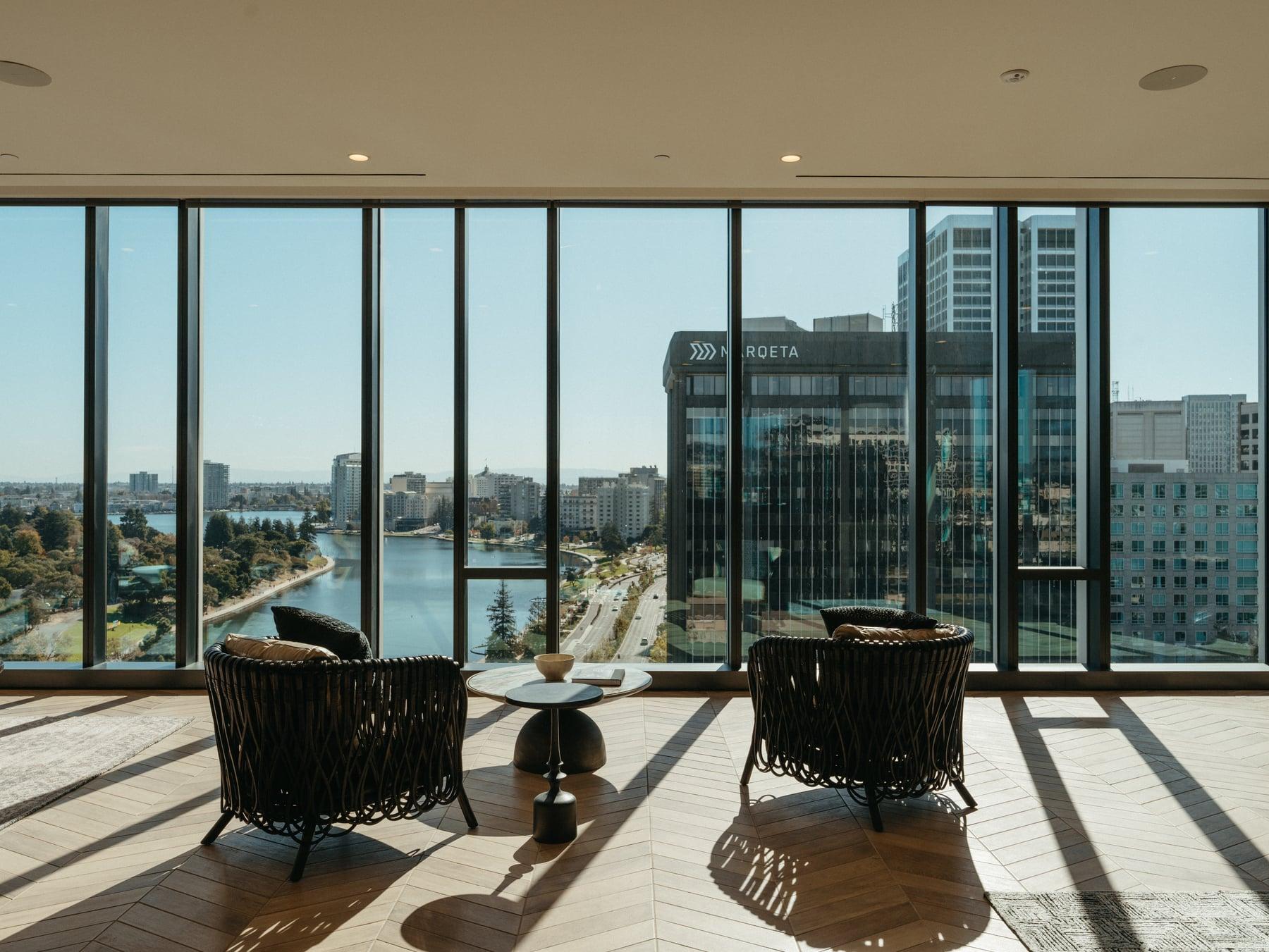 Interior lounge with floor-to-ceiling windows, two woven chairs and a view of water and city buildings