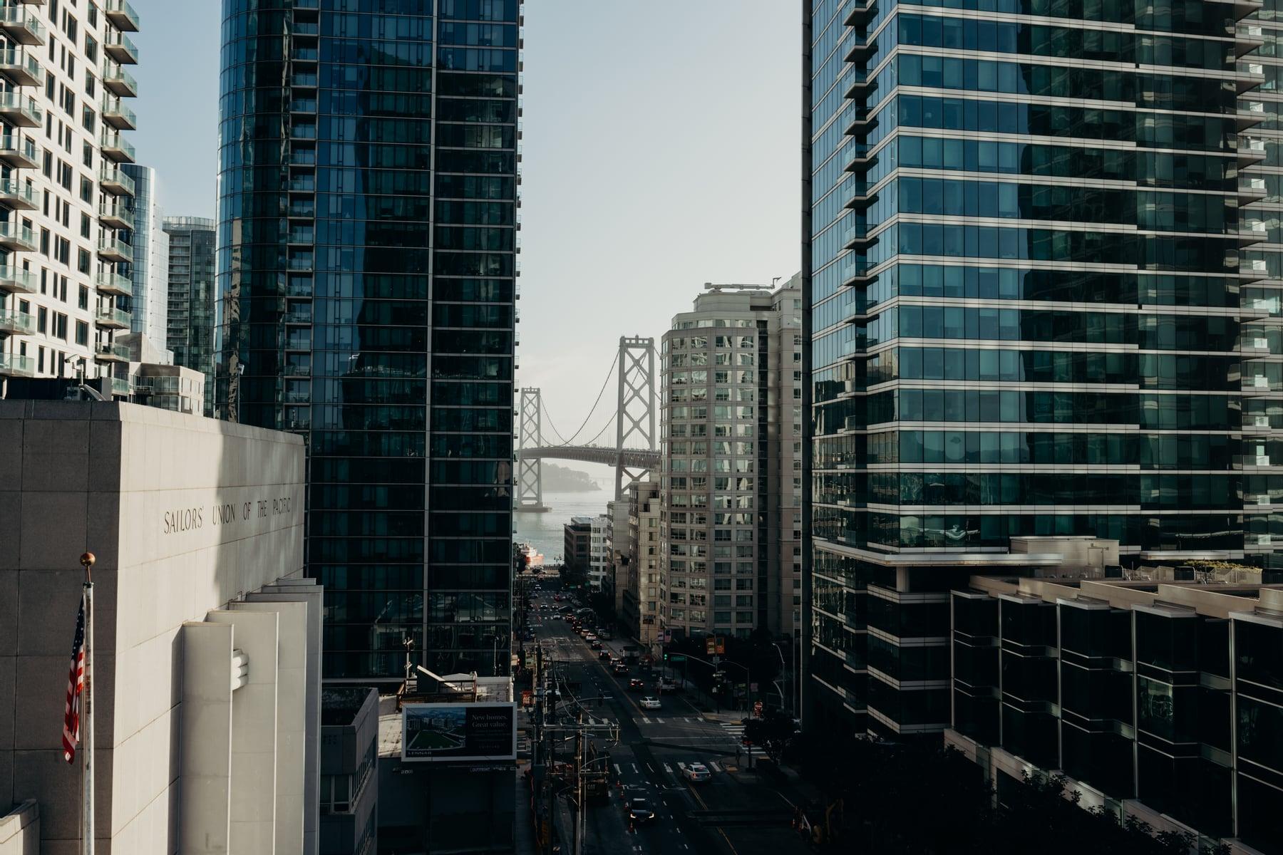 View down a San Francisco street between glass towers, with a bridge over the bay in the distance