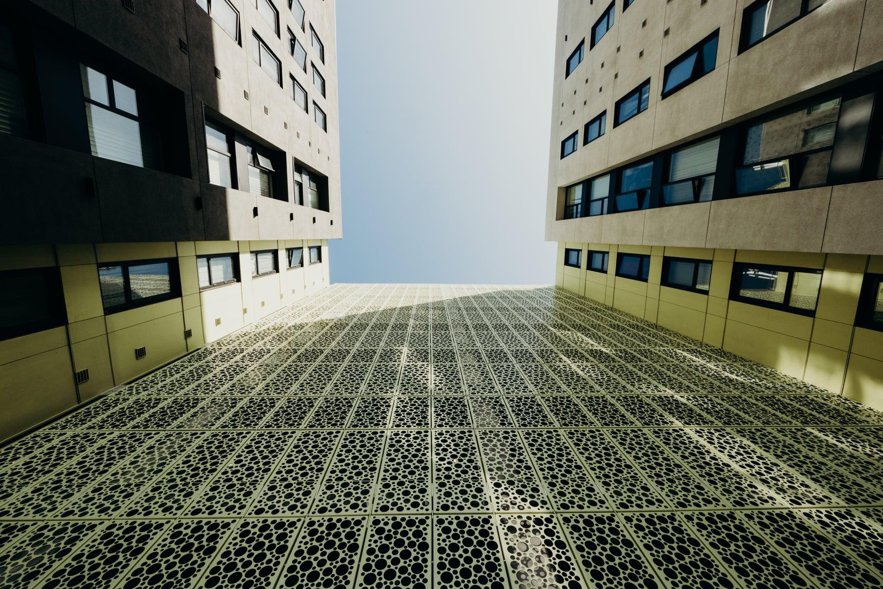Looking up between two buildings over a courtyard paved with a perforated pattern, with yellow-green paneled walls