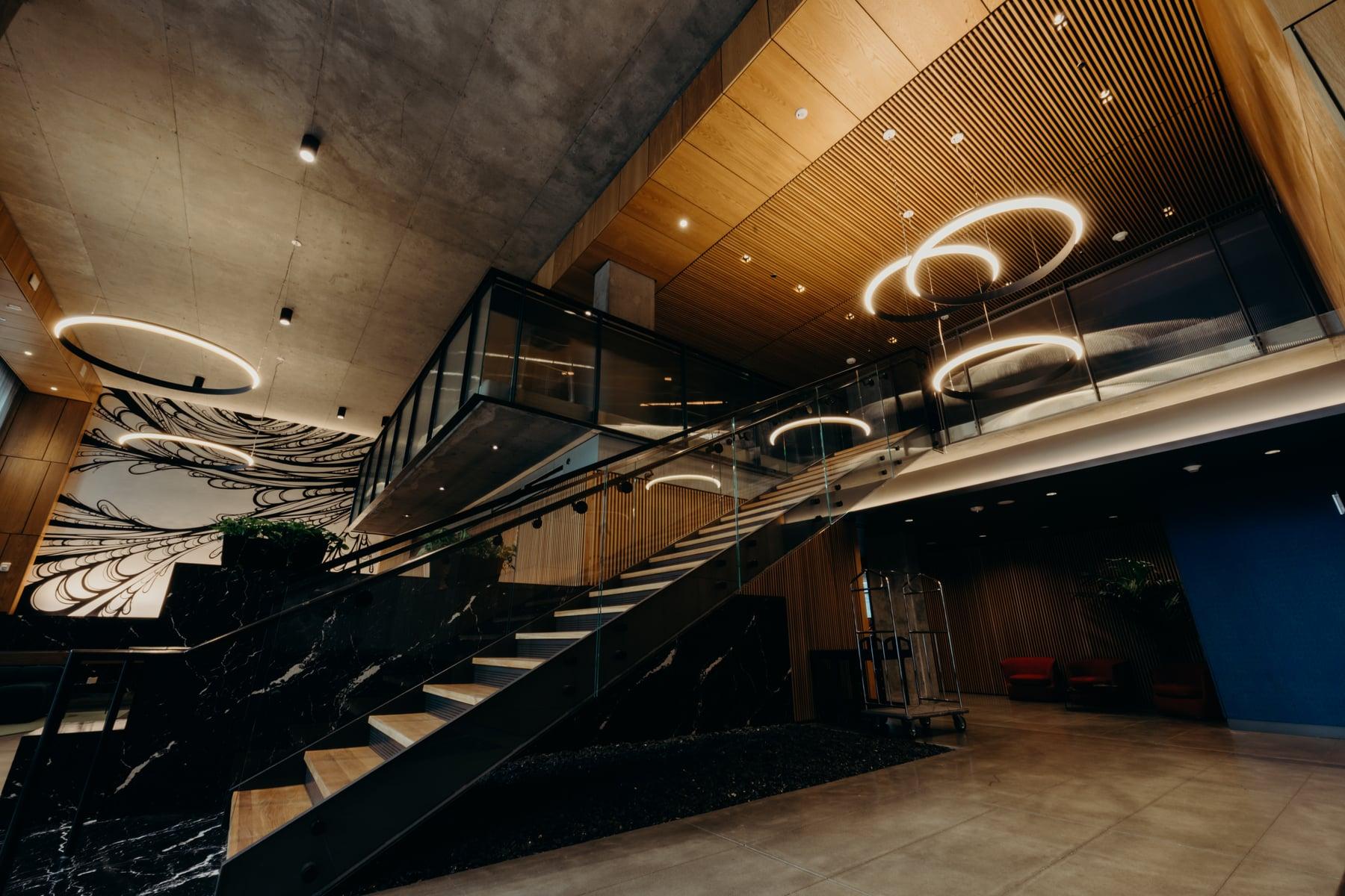 Lobby with a dark stone feature wall, an open staircase with glass guardrail and a wood-slat ceiling with ring-shaped lights