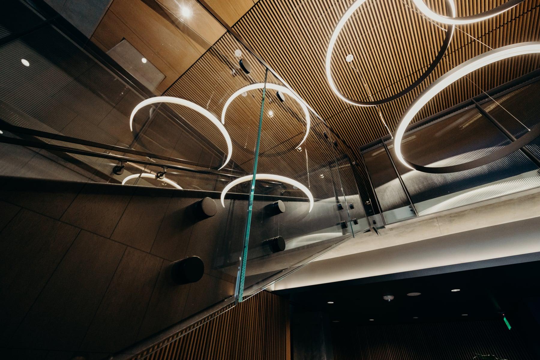 Glass guardrail along the mezzanine beneath circular light fixtures and a wood-slat ceiling