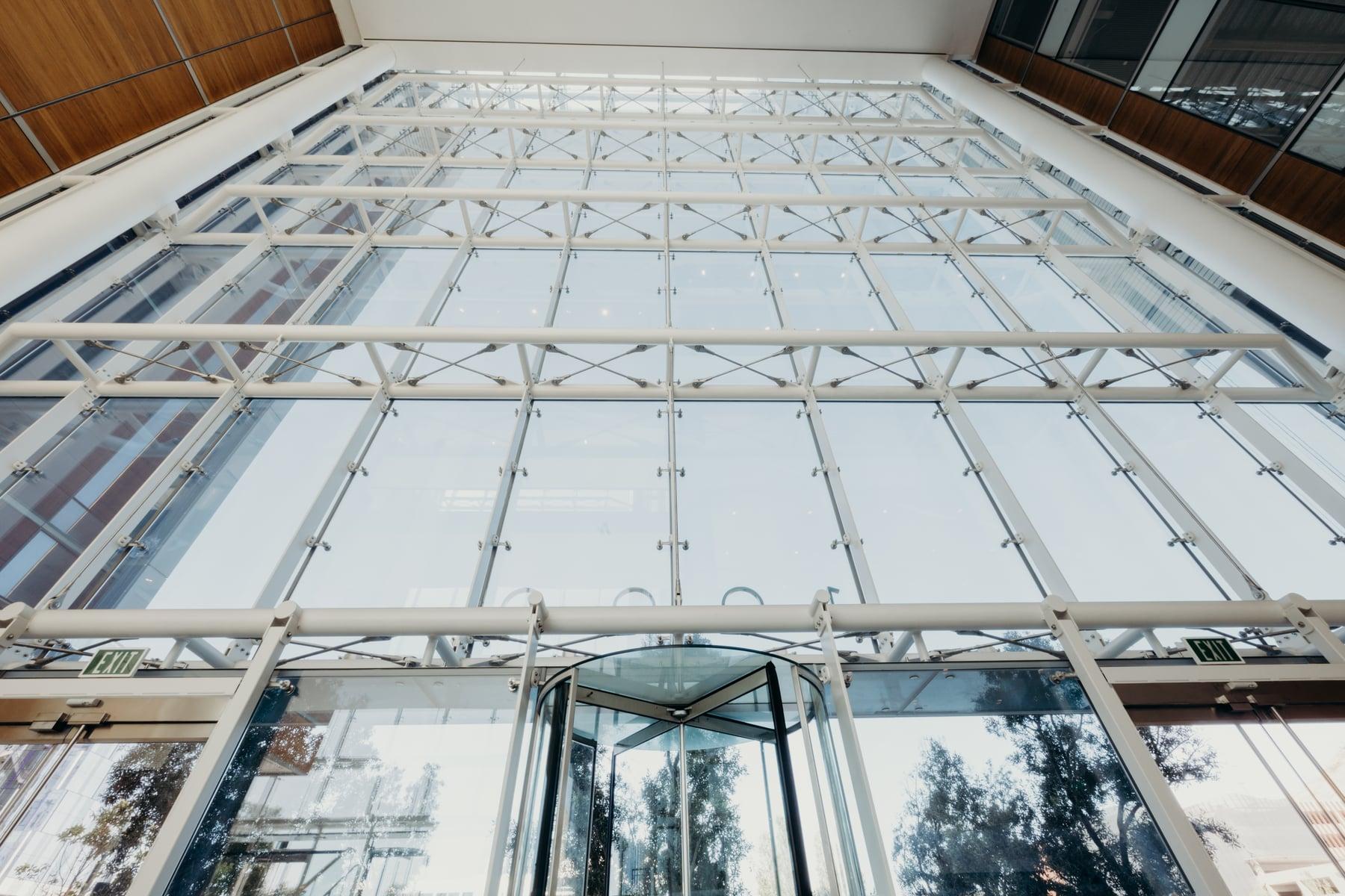 Interior looking up at a tall glass wall held by a white steel frame above a revolving door