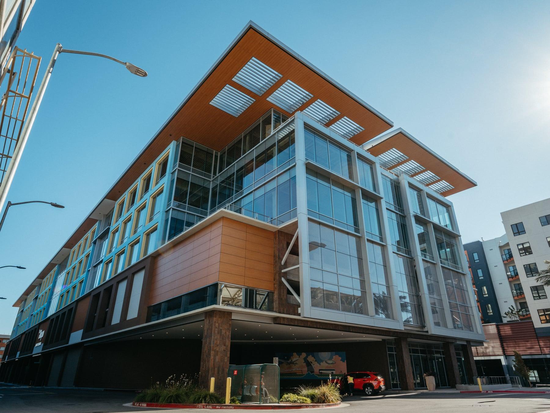 Corner of the Millbrae mixed-use building with a wood-toned roof overhang, glazed corner bays and a car parked at street level
