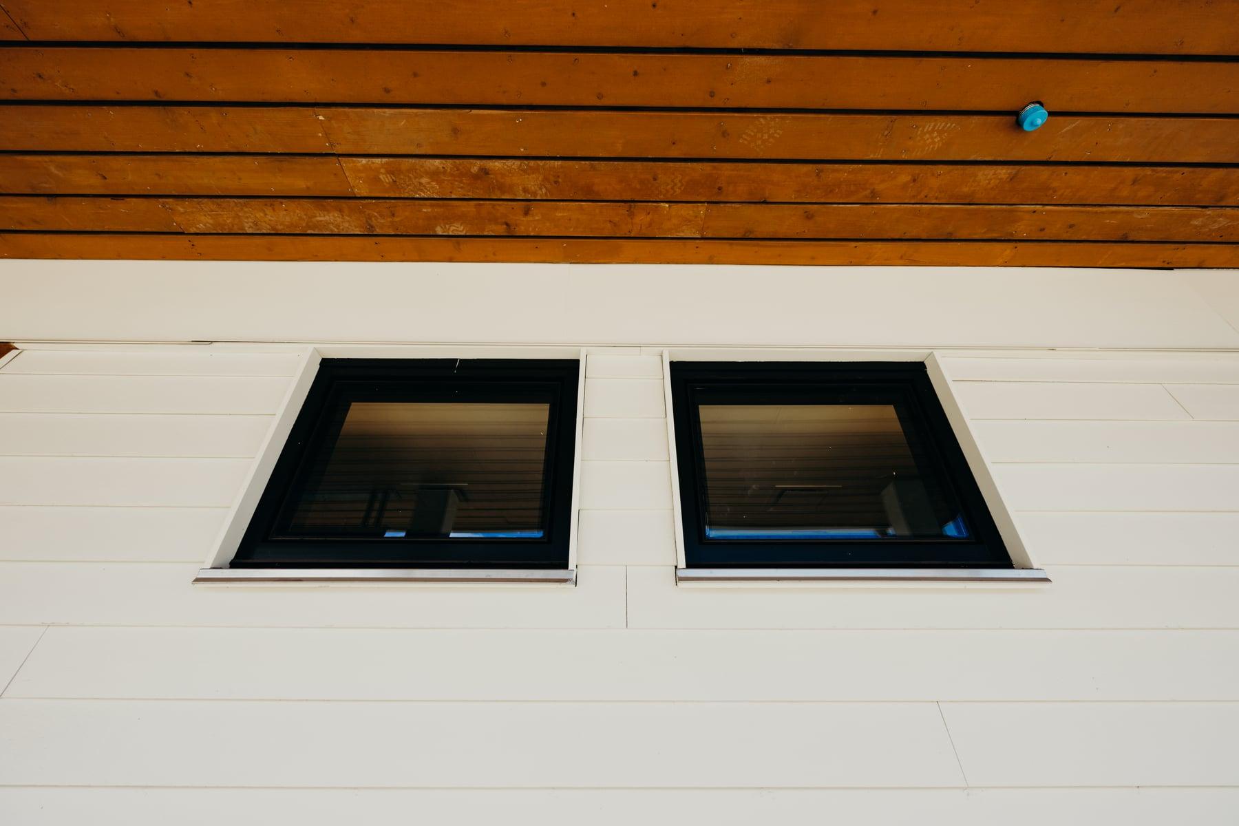 Two small black-framed windows set in white siding below a wood-slat soffit