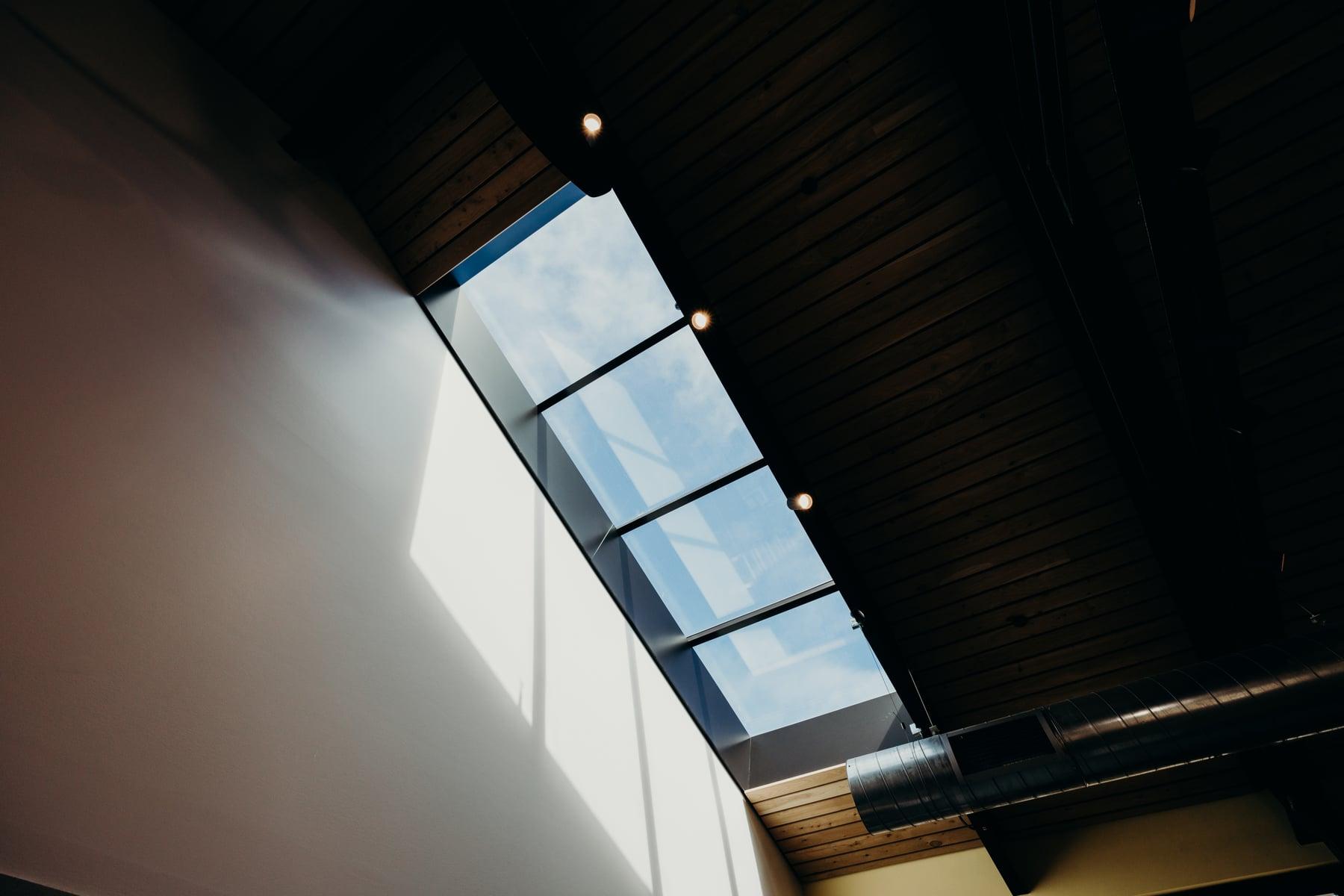 Skylight slanting along a dark wood-lined ceiling and white wall, with sky visible through the glass