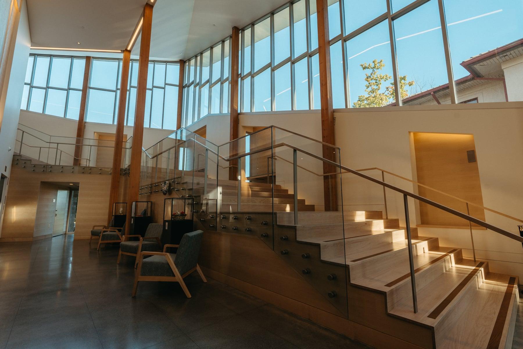 Interior stair with a glass and steel handrail and lit steps, beside a tall glazed wall and wood columns