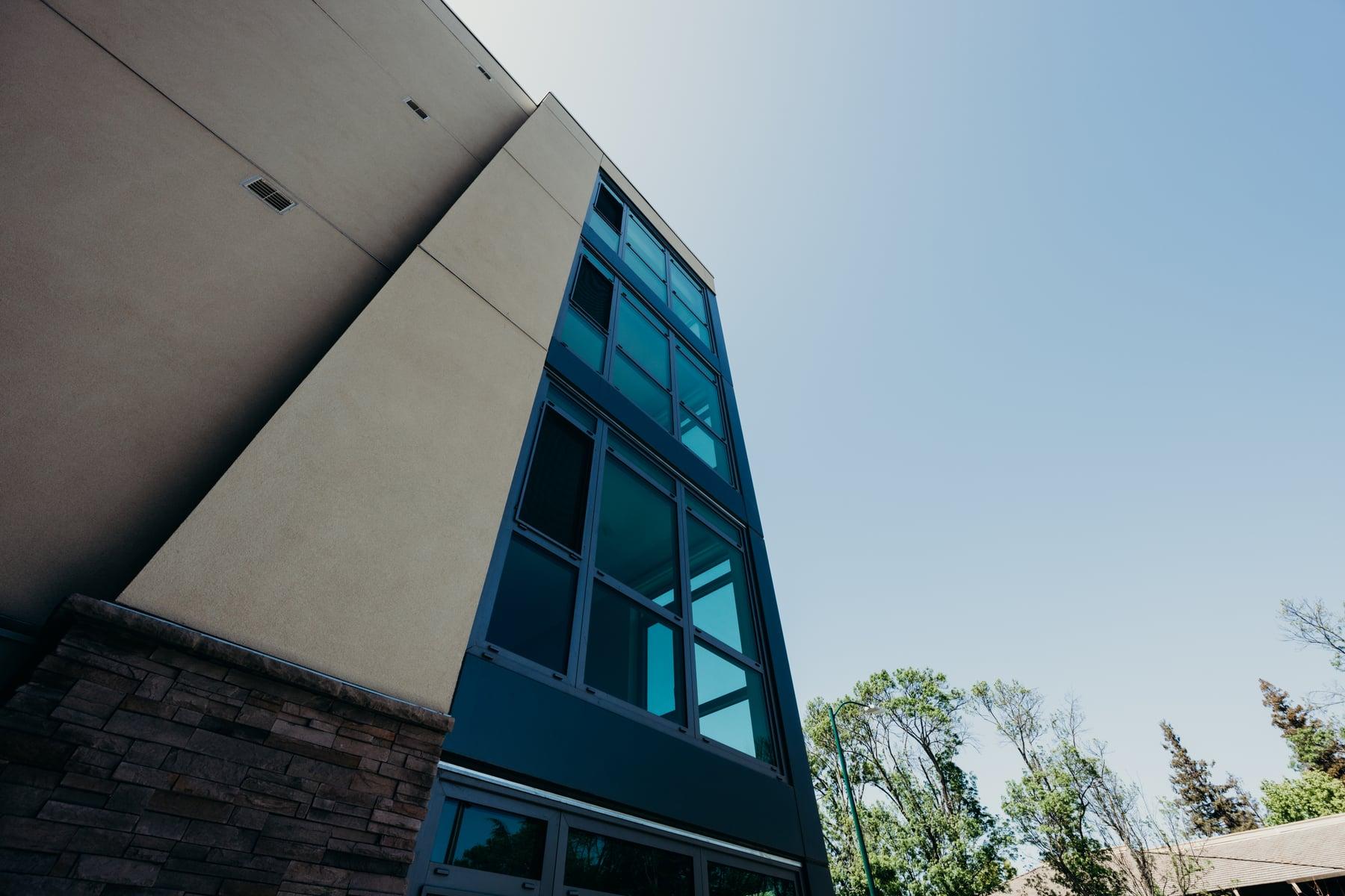 Looking up at a tall dark-framed glazed bay beside a stone-clad base and a stucco wall