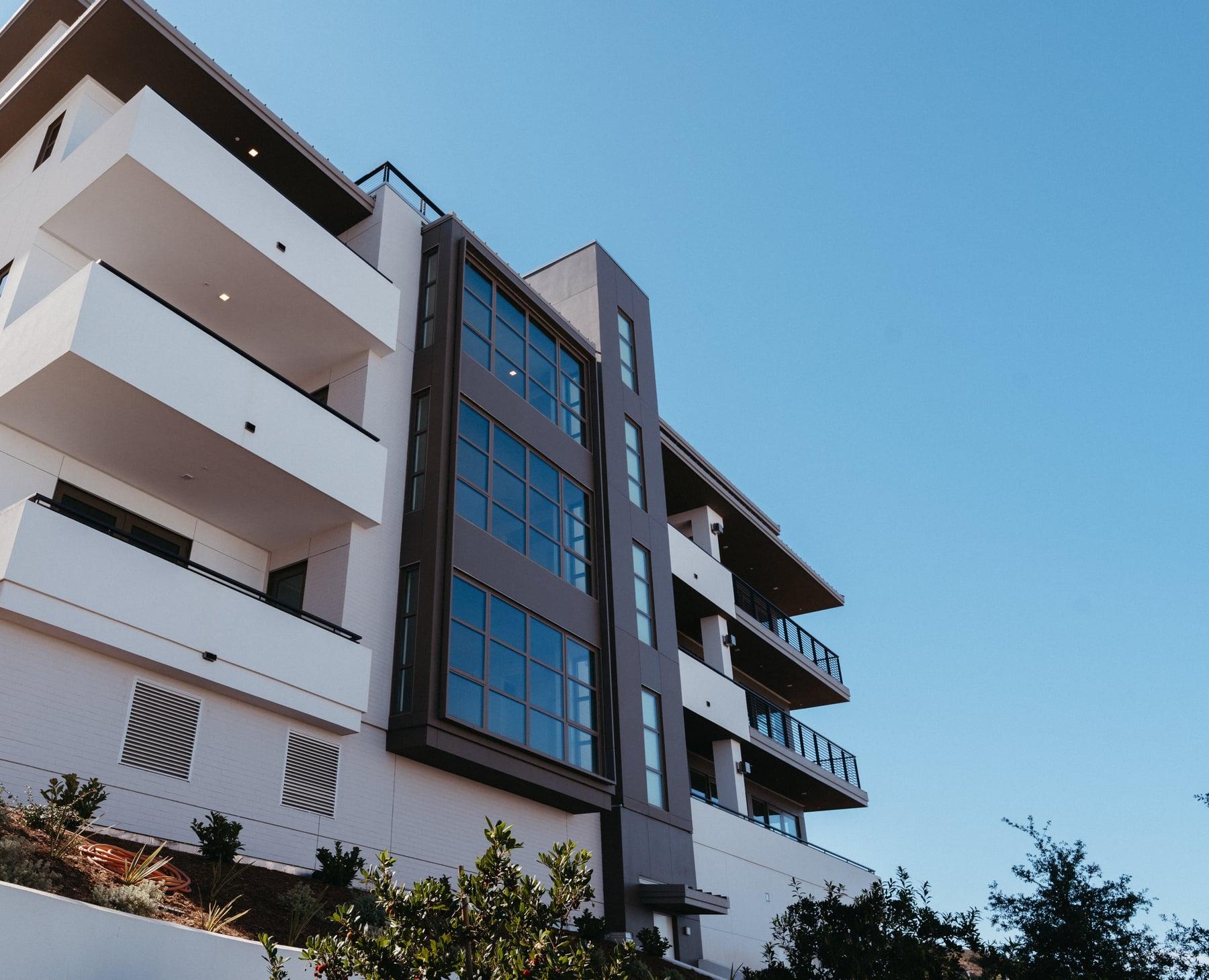 Looking up at a residential building with white balconies and a tall dark-framed glazed bay