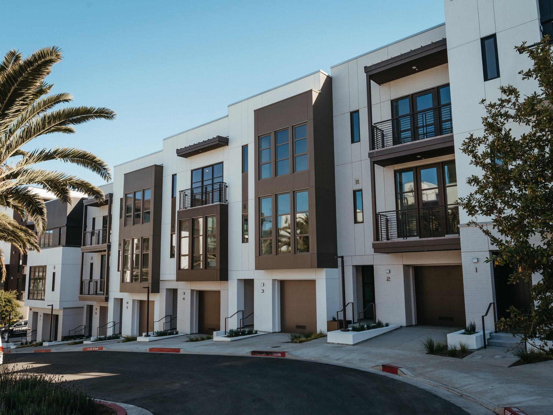 Row of white and dark-paneled townhomes with garages, windows above and a palm tree along a curving street