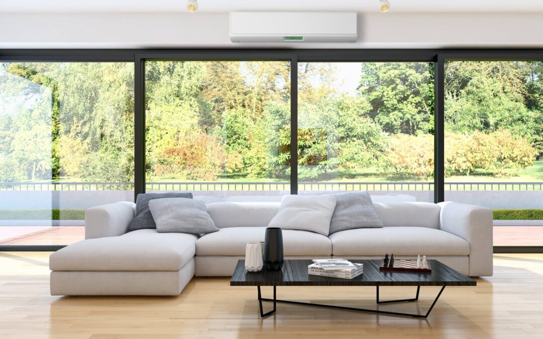 A modern living room showcasing the latest interior glazing trends with floor-to-ceiling glass windows that provide a seamless connection to the lush green outdoor view. The space features a sleek white sectional sofa with gray throw pillows, a black coffee table with decorative items, including a chessboard, on a light wood floor, and an air conditioning unit mounted on the wall above, enhancing the clean and minimalist aesthetic.